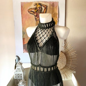 May include: A long, olive green crochet dress with a halter neckline and fringe detailing. The dress has a geometric pattern and a fitted bodice.