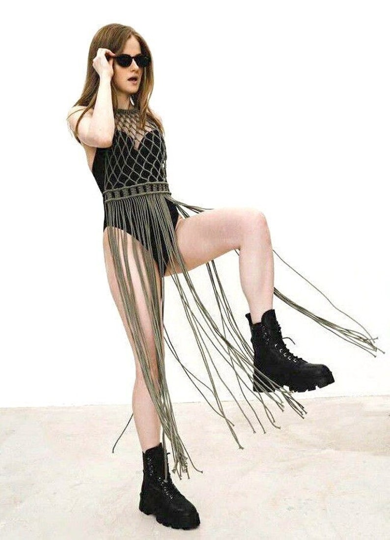 May include: A woman wearing a black bodysuit with a green crochet fringe top and black boots. She is wearing sunglasses and has her hair down.