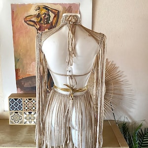 May include: A beige macrame top featuring long fringe sleeves and a halter neck design. The garment includes a high neck collar, an open back with tie closures, and a metallic gold waist tie. The piece is constructed with knotted cord.