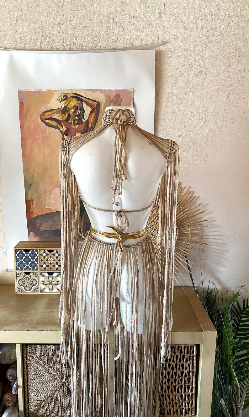 May include: A beige macrame top featuring long fringe sleeves and a halter neck design. The garment includes a high neck collar, an open back with tie closures, and a metallic gold waist tie. The piece is constructed with knotted cord.