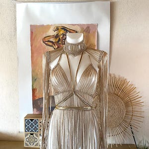 May include: A beige macrame garment featuring a high-neck choker collar, long fringe sleeves, and a bodice with thin straps. The piece is constructed with intricate knotted cord detailing, creating a textured, layered appearance that drapes down.