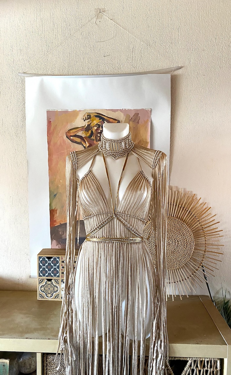 May include: A beige macrame garment featuring a high-neck choker collar, long fringe sleeves, and a bodice with thin straps. The piece is constructed with intricate knotted cord detailing, creating a textured, layered appearance that drapes down.