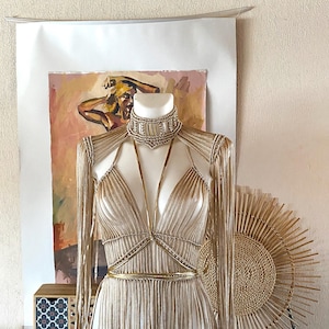 May include: A beige macrame top featuring a high neck collar, long fringe sleeves, and a deep V-neckline. The garment is accented with thin, metallic gold straps that cross over the chest and wrap around the waist, creating a structured, layered appearance.