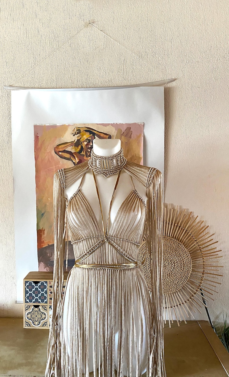 May include: A beige macrame top featuring a high neck collar, long fringe sleeves, and a deep V-neckline. The garment is accented with thin, metallic gold straps that cross over the chest and wrap around the waist, creating a structured, layered appearance.