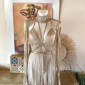 May include: A beige macrame dress featuring a high-neck choker collar, a deep V-neckline, and long sleeves. The garment is constructed with intricate knotted cord patterns and long, flowing fringe details that extend from the shoulders to the hem.