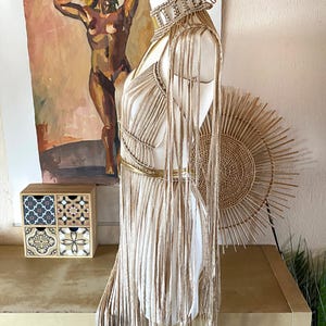 May include: A beige macrame dress featuring a high-neck choker collar, long fringe detailing extending from the shoulders, and a metallic gold waist belt. The garment is constructed with intricate knotted cord patterns and long, flowing vertical strands.