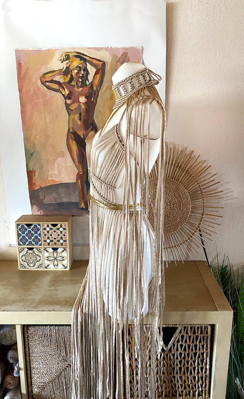 May include: A beige macrame dress featuring a high-neck choker collar, long fringe detailing extending from the shoulders, and a metallic gold waist belt. The garment is constructed with intricate knotted cord patterns and long, flowing vertical strands.