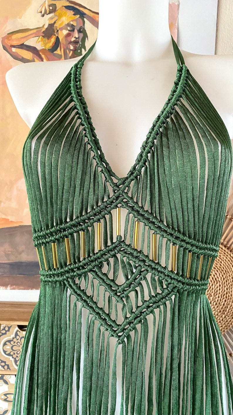 Op de afbeelding: Een groene gecrochete haltertop met gouden kralen en franjes. De top is gemaakt met een macram&eacute;-steek en heeft een V-hals.
