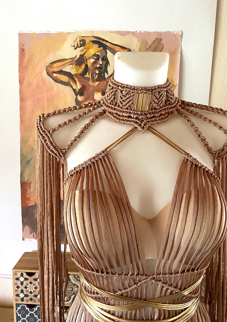May include: A copper-colored macrame top featuring a high-neck choker collar, intricate knotted shoulder detailing, and long fringe sleeves. The garment is constructed with vertical cord panels and a layered metallic gold waist wrap over a beige base.