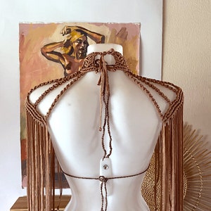 May include: A light brown macrame halter top featuring an intricate knotted neck design, multiple shoulder straps, and long, flowing fringe detailing that extends down the arms and back, secured with thin adjustable ties at the neck and mid-back.