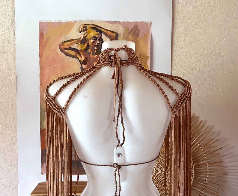 May include: A light brown macrame halter top featuring an intricate knotted neck design, multiple shoulder straps, and long, flowing fringe detailing that extends down the arms and back, secured with thin adjustable ties at the neck and mid-back.