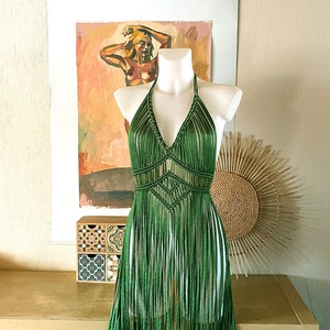 Op de afbeelding: Een groene gehaakte haltertop met franjes. De top is op een wit mannequin en wordt getoond tegen een lichtbruine muur.