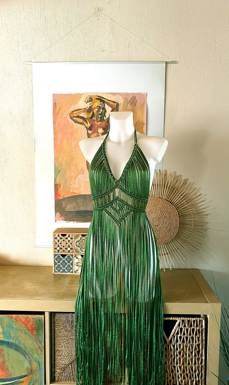 Op de afbeelding: Een groene gehaakte haltertop met franjes. De top is op een wit mannequin en wordt getoond tegen een lichtbruine muur.