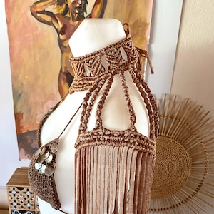 May include: A brown macrame halter top featuring a high neck collar, intricate knotted shoulder straps, and long fringe detailing. The garment is embellished with small, circular, iridescent shell accents on the front triangular bodice section.