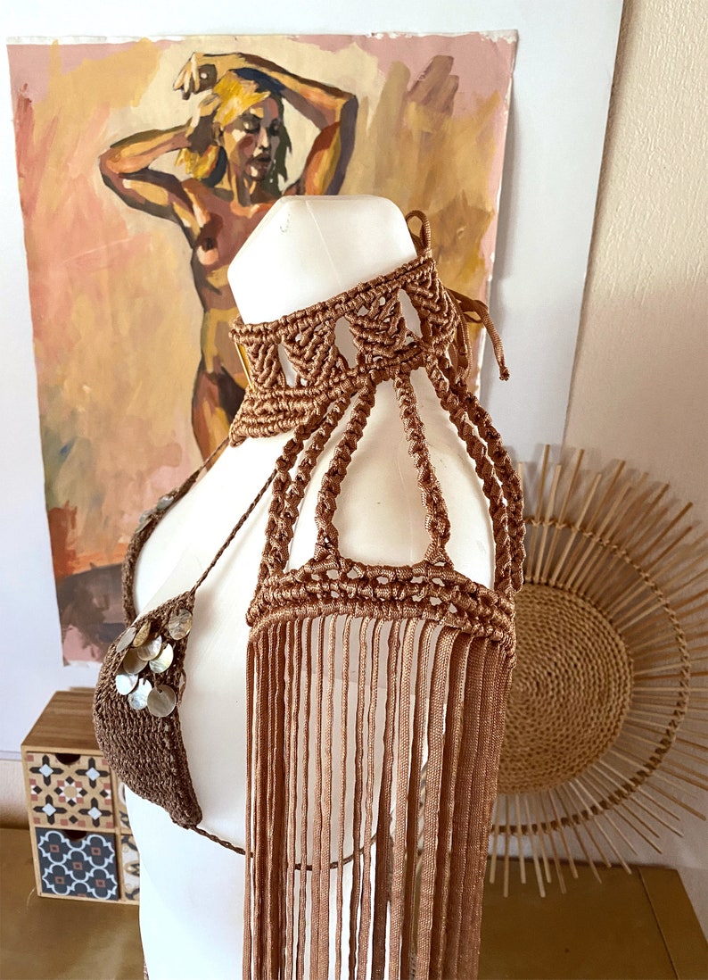May include: A brown macrame halter top featuring a high neck collar, intricate knotted shoulder straps, and long fringe detailing. The garment is embellished with small, circular, iridescent shell accents on the front triangular bodice section.