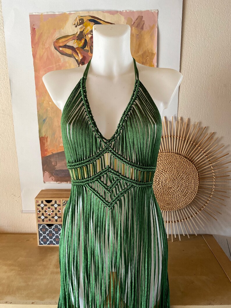 Op de afbeelding: Een groene gehaakte haltertop met gouden kralen en franjes. De top is op een wit mannequin.