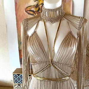 May include: A beige macrame dress featuring a high-neck choker collar with vertical accents, long fringe sleeves, and a deep V-neckline. The garment is accented with thin, metallic gold straps that cross at the chest and wrap around the waist.