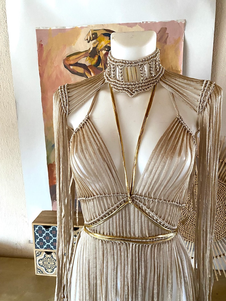 May include: A beige macrame dress featuring a high-neck choker collar with vertical accents, long fringe sleeves, and a deep V-neckline. The garment is accented with thin, metallic gold straps that cross at the chest and wrap around the waist.