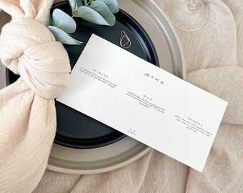 May include: A white menu card with a black background. The menu card is divided into three sections: Starter, Main, and Dessert. The text on the menu card is in a simple, elegant font. The menu card is placed on a beige tablecloth with a white napkin and a sprig of eucalyptus.