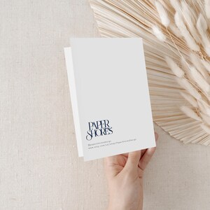 Puede incluir: Una tarjeta plegada blanca con el texto "PAPER SHORES" en una fuente de escritura azul oscuro. La tarjeta est&aacute; sostenida por una mano, con flores secas y un fondo beige.