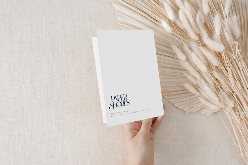 Puede incluir: Una tarjeta plegada blanca con el texto "PAPER SHORES" en una fuente de escritura azul oscuro. La tarjeta est&aacute; sostenida por una mano, con flores secas y un fondo beige.