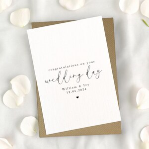 May include: A white card with black text that reads "congratulations on your wedding day" with the names "William & Ivy" and the date "12.05.2024" below. The card is sitting on a brown envelope.