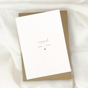 Pode incluir: Um cart&atilde;o branco com a palavra "engaged" escrita em it&aacute;lico. Abaixo da palavra, diz "Jake + Lois" em vermelho com um pequeno cora&ccedil;&atilde;o vermelho.
