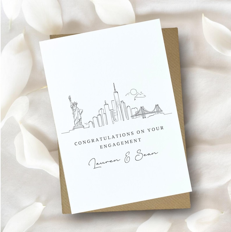 Op de afbeelding: Een witte kaart met een zwarte lijntekening van de skyline van New York City, inclusief het Vrijheidsbeeld en de Brooklyn Bridge. De tekst "Congratulations on your engagement" en "Lauren & Sean" staat onder de tekening.