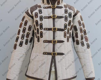 May include: A cream-colored quilted jacket with brown leather accents and gold buckles. The jacket features a high collar and is designed with rectangular leather patches on the sleeves and front. The jacket is part of a medieval or historical clothing collection.