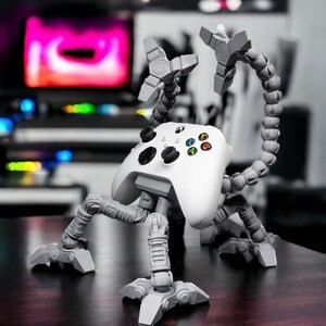 May include: A white Xbox controller with a 3D printed, articulated, gray stand that resembles a spider. The stand has multiple joints and legs, allowing the controller to be positioned in various poses.