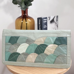 Peut inclure: Une pochette en cuir vert clair avec un motif patchwork de demi-cercles superposés dans des tons de vert, de gris et de beige. La pochette est rectangulaire avec une surface texturée et une bordure vert clair. Elle est posée sur une table en bois.