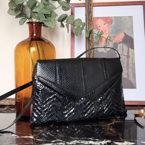 Può includere: Borsa a tracolla in pelle nera con motivo a zig-zag sul davanti. La borsa ha una tracolla lunga e sottile e una forma rettangolare. Un vaso con foglie verdi e un dipinto incorniciato sono sullo sfondo.