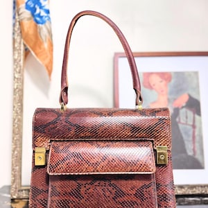 Peut inclure: Un sac à main vintage en peau de serpent marron avec une poignée supérieure et une poche à rabat avant. Le sac a une quincaillerie dorée et une forme rectangulaire. La poignée et la garniture sont marron foncé. Un foulard et une œuvre encadrée sont en arrière-plan.