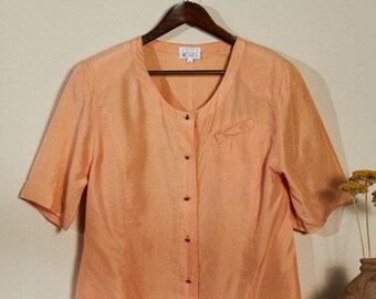 Puede incluir: Una blusa de color melocotón con botones y mangas cortas. La blusa tiene un escote redondo y un solo bolsillo en el lado izquierdo. La etiqueta de la marca dice "Oui Korn".