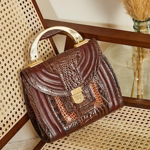 Peut inclure: Sac à main vintage en cuir marron avec fermoir et poignée dorés. Le sac présente un motif texturé avec un mélange de tons marron foncé et clair. Le sac est posé sur une chaise en osier.