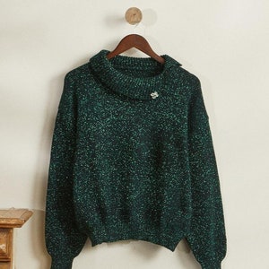 Può includere: Un maglione verde scuro con collo a imbuto e maniche lunghe, macchiato di fili verde chiaro. Una piccola spilla decorativa è appuntata vicino allo scollo. Il maglione è appeso a una gruccia di legno.