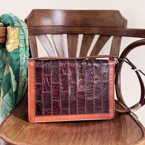 May include: A vintage handbag with a patchwork design. The bag has a rectangular shape with a flap closure. The main body is a deep burgundy colour, with a textured pattern, and is framed by a lighter brown border. A long strap is attached.