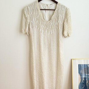 May include: A white beaded dress with a V-neckline and short sleeves. The dress has a scalloped hem and is made of a sheer fabric.