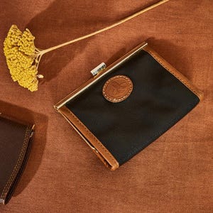 Könnte beinhalten: Eine schwarze Lederbörse mit braunem Lederbesatz und einem goldenen Metallverschluss. Die Börse hat einen kleinen, runden, braunen Lederfleck mit einem Logo auf der Vorderseite.