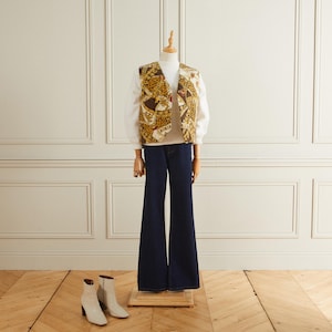 Puede incluir: Un chaleco estampado en marrón y dorado con una camisa blanca de manga larga debajo. La modelo lleva un vaquero azul oscuro acampanado y botas blancas de tacón.