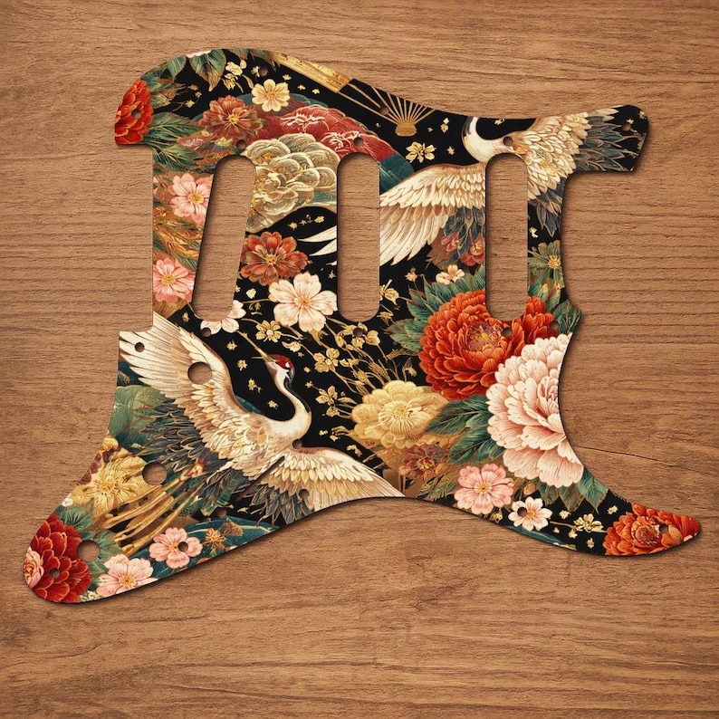 Puede incluir: Un golpeador de guitarra con un dise&ntilde;o de inspiraci&oacute;n japonesa. El golpeador presenta un fondo negro con un patr&oacute;n floral, que incluye flores rojas, rosas y amarillas, e im&aacute;genes de grullas. El golpeador tiene tres agujeros para pastillas.