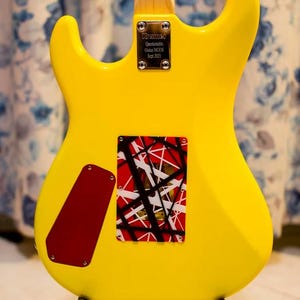 May include: A bright yellow electric guitar, viewed from the back. It features a red and black patterned cover and a red cover plate. The guitar has a black stand and a neck with a wooden finish. The text "Kramer Questionable Guitar MODS Sept 2021" is visible.