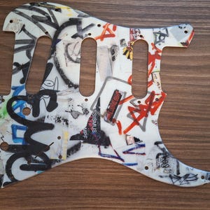 May include: White guitar pickguard with a graffiti design featuring black, red, blue, and yellow spray paint. The pickguard has three pickup cutouts and several screw holes. The design includes various words and symbols.