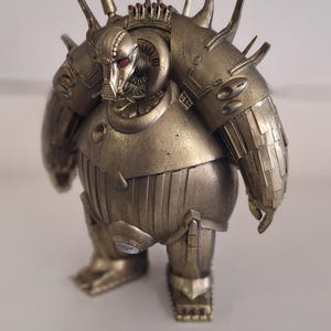 Peut inclure: Une figurine de robot de couleur dorée avec un design détaillé et piquant. Le robot a un corps rond et une petite tête avec des yeux rouges.