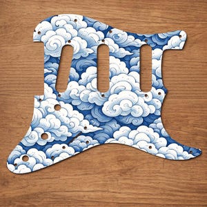 Puede incluir: Un golpeador de guitarra con un dise&ntilde;o de nubes azules y blancas. El golpeador tiene tres cortes para pastillas y varios agujeros para tornillos. El dise&ntilde;o presenta nubes arremolinadas en tonos azules y blancos.