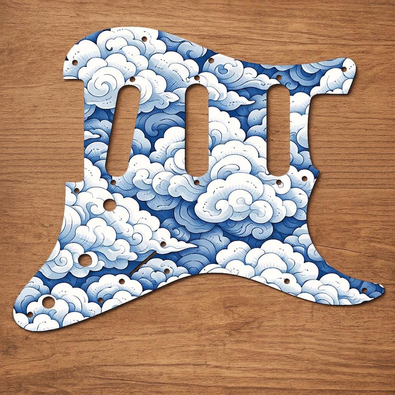 Puede incluir: Un golpeador de guitarra con un dise&ntilde;o de nubes azules y blancas. El golpeador tiene tres cortes para pastillas y varios agujeros para tornillos. El dise&ntilde;o presenta nubes arremolinadas en tonos azules y blancos.
