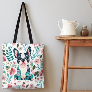 Puede incluir: Una bolsa de tela blanca con asa negra presenta una ilustraci&oacute;n colorida de un bulldog franc&eacute;s con motivos florales. El fondo es un lienzo blanco con dise&ntilde;os florales rosas, azules y verdes.