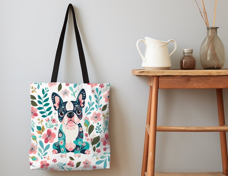 Puede incluir: Una bolsa de tela blanca con asa negra presenta una ilustraci&oacute;n colorida de un bulldog franc&eacute;s con motivos florales. El fondo es un lienzo blanco con dise&ntilde;os florales rosas, azules y verdes.