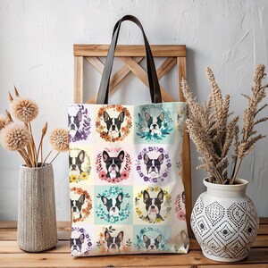 K&ouml;nnte beinhalten: Eine cremefarbene Tragetasche mit einem sich wiederholenden Muster von Boston Terrier-Hundeportr&auml;ts in Blumenkr&auml;nzen. Sie hat braune Ledergriffe. Die Tasche wird auf einer Holzoberfl&auml;che pr&auml;sentiert. Der Hintergrund umfasst dekorative Vasen mit Trockenblumen.