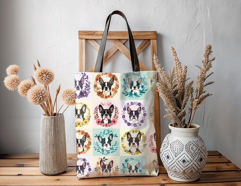 K&ouml;nnte beinhalten: Eine cremefarbene Tragetasche mit einem sich wiederholenden Muster von Boston Terrier-Hundeportr&auml;ts in Blumenkr&auml;nzen. Sie hat braune Ledergriffe. Die Tasche wird auf einer Holzoberfl&auml;che pr&auml;sentiert. Der Hintergrund umfasst dekorative Vasen mit Trockenblumen.
